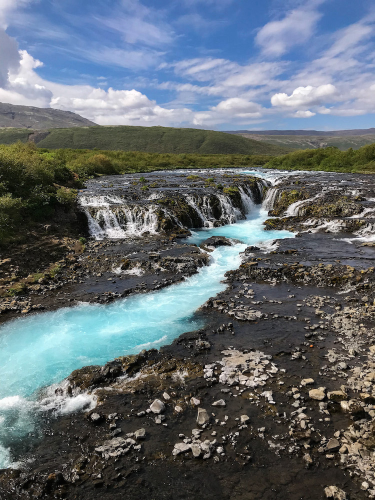 Der Bruarfoss in Island beginnt weit und flach auf schwarzem Untergrund und ergibt sich in der Mitte schmal über die dunklen Steine und formiert darunter einen breiten türkisfarbenen Flusslauf.