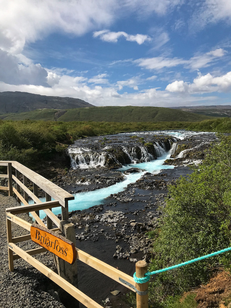 Der Bruarfoss in Island beginnt weit und flach auf schwarzem Untergrund und ergibt sich in der Mitte schmal über die dunklen Steine und formiert darunter einen breiten türkisfarbenen Flusslauf.