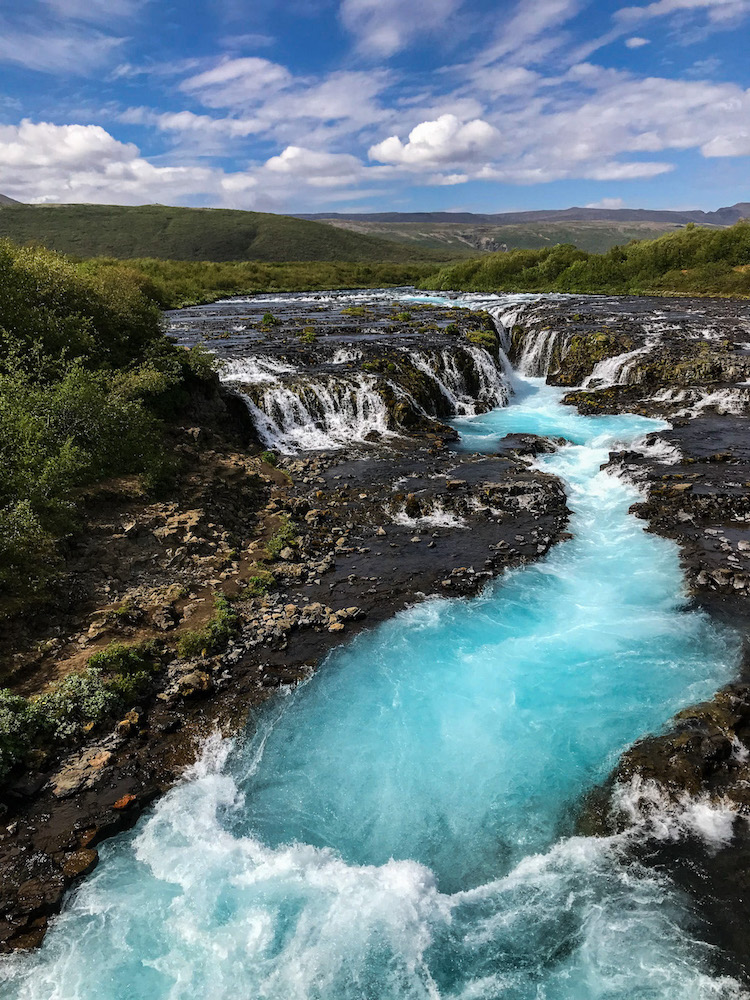 Der Bruarfoss in Island beginnt weit und flach auf schwarzem Untergrund und ergibt sich in der Mitte schmal über die dunklen Steine und formiert darunter einen breiten türkisfarbenen Flusslauf.