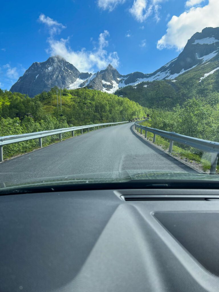 Ausblick aus der Frontscheibe eines Auto auf eine steile Bergstraße, schneebedeckte Gipfel voraus.