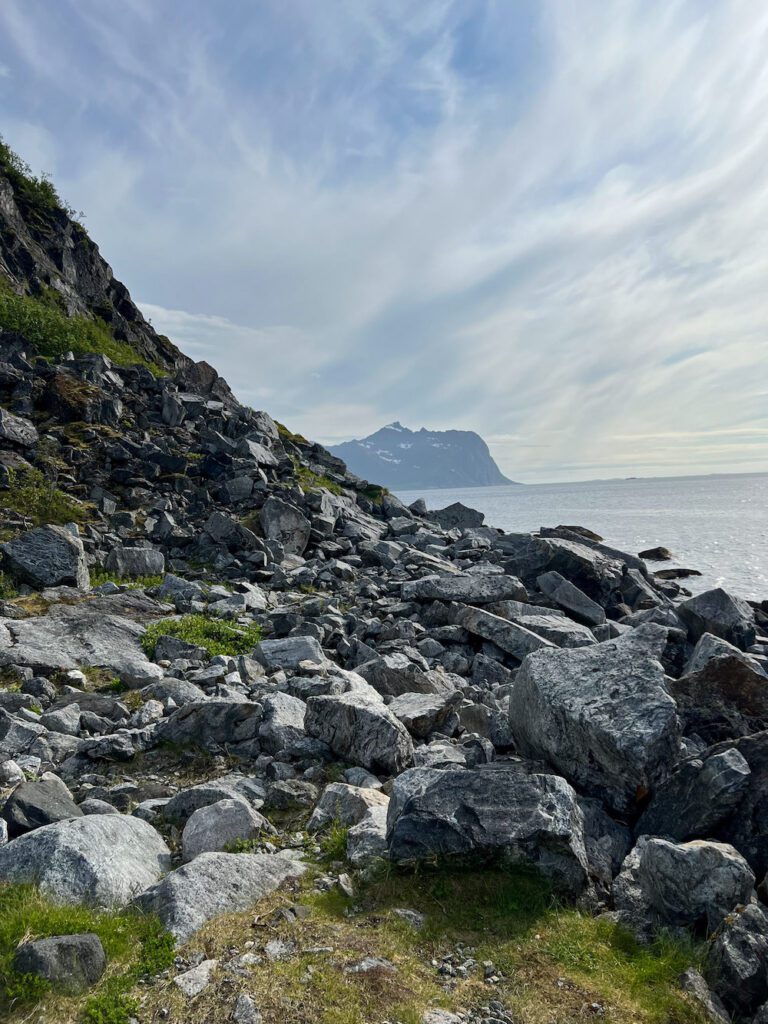 große Steine an einem Hang, dahinter das Meer und ein hoher Berg am Horizont