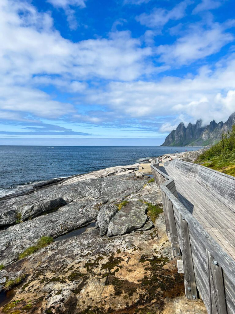 Holzsteg über steinigem Untergrund führt entlang des Fjords zum Ausblick auf Berggipfel