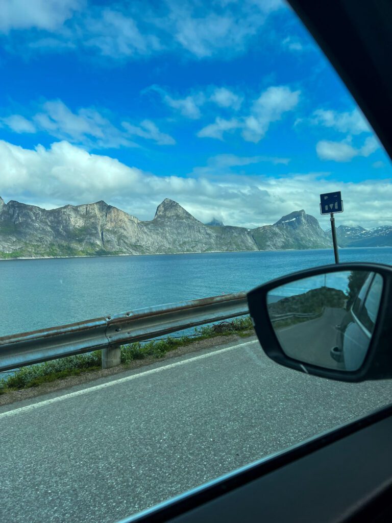 Ausblick aus Autofenster auf Strasse und Fjord dahinter, hohe Bergkette am Horizont