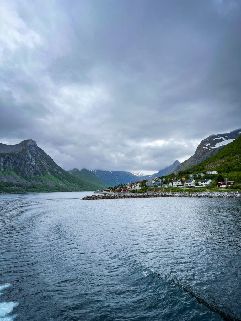 Blick in den Fjord von der Fähre aus, Gryllefjord mit den bunten Häusern am rechten Ufer, dunkle Wolken über den Bergen