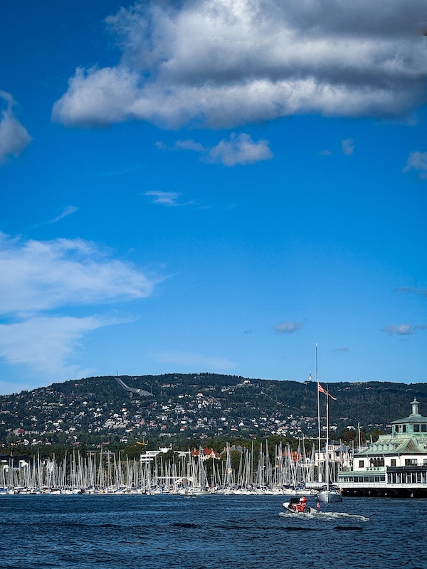 Jachthafen von Oslo vorne im Bild, hinten die Stadt, die sich hoch an den Berg schmiegt bis hinauf zur Schanze auf dem Holmenkollen vor blauen Himmel